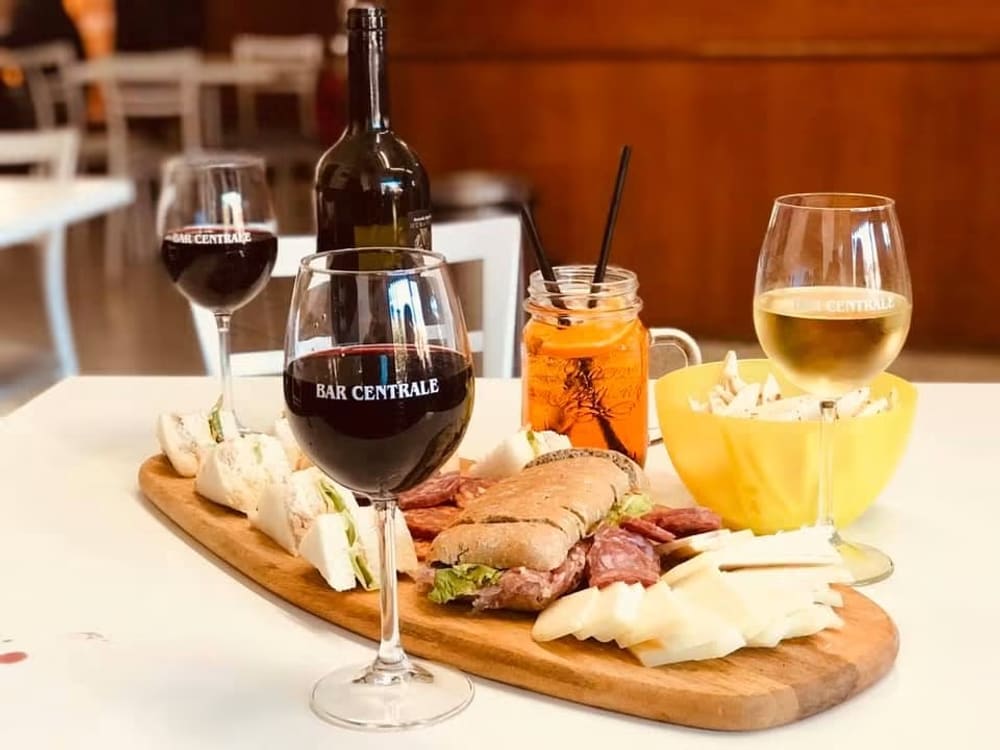 Tagliere di salumi, formaggi e rotolini di piadina su un tavolo del Bar Centrale, con una bottiglia di vino rosso e i calici serviti